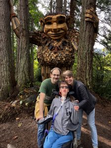 Burke, Lucas, Krista in front of large wooden troll sculpture in the woods.